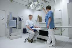 Patient sitzt vor Röntgen-Liege mit Unterarm auf der Liege MTRA steht vor ihm und richtet das an der Decke hängende Röntgengerät über dem Arm aus.