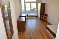 Bild möbliertes Zimmer links Garderobe, Sideboard und Schreibtisch mit Stuhl rechts Bett Stuhl und Kleiderschrank zimmerbreites bodentiefes Fenster Laminatboden