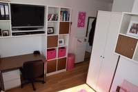 Blick vom Bett ins Zimmer. An Wand zum Bad Kleiderschrank und Regal über Fußende Bett. Linke Wand Schreibtisch und umlaufendes Regal mit TV und kleinen Schränken. Laminatboden. Dekorationen in Pink